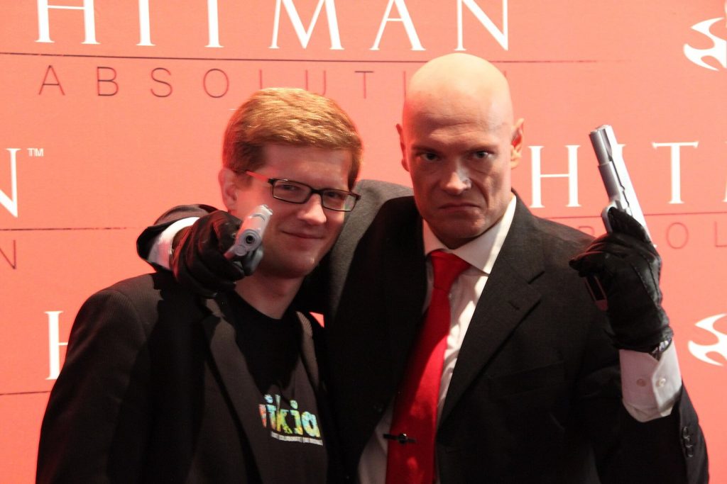 Model dressed as Agent 47 from Hitman posing with a fan at Gamescom 2012